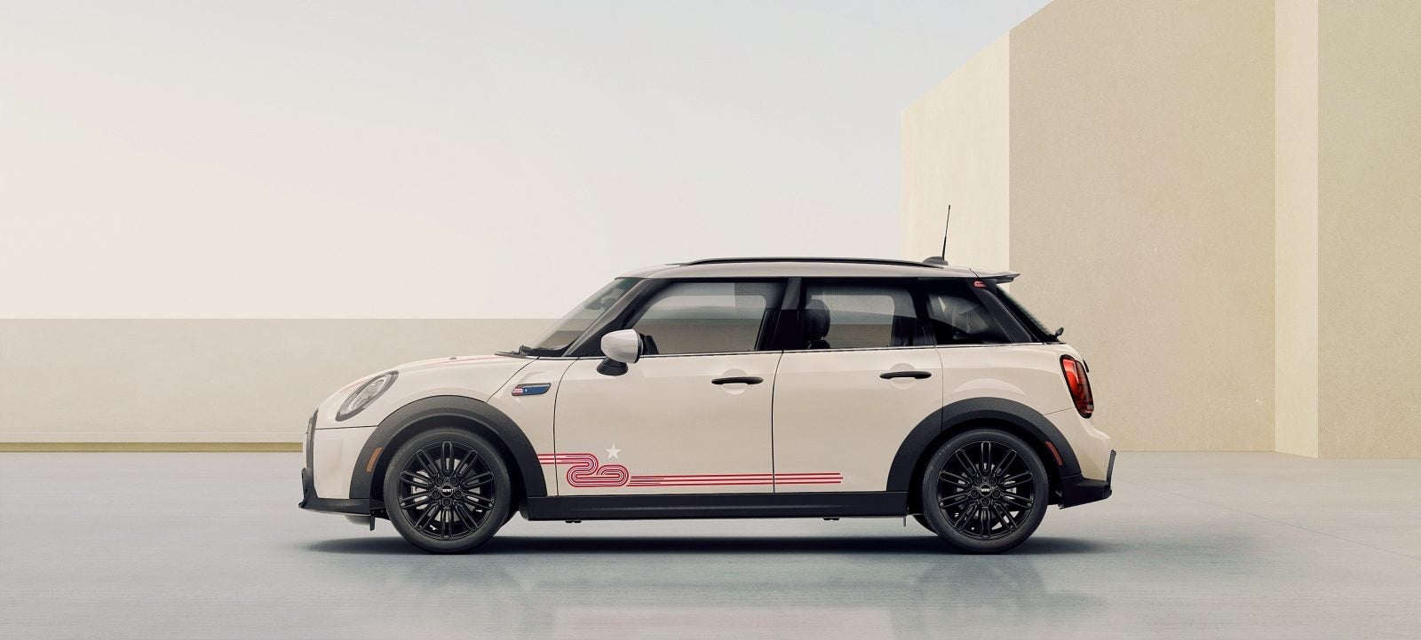 Vista lateral de un MINI 20 Years Edition en Pepper White mirando hacia la izquierda, aparcado en una superficie gris con edificios rectangulares beige y un cielo despejado al fondo. | MINI of Sterling en Sterling VA