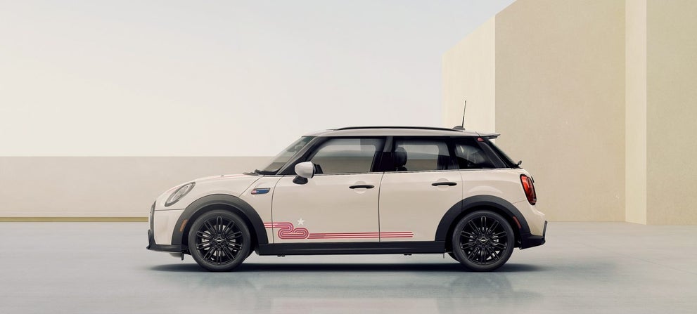 Vista lateral de un MINI 20 Years Edition en Pepper White mirando hacia la izquierda, aparcado en una superficie gris con edificios rectangulares beige y un cielo despejado al fondo. | MINI of Sterling en Sterling VA