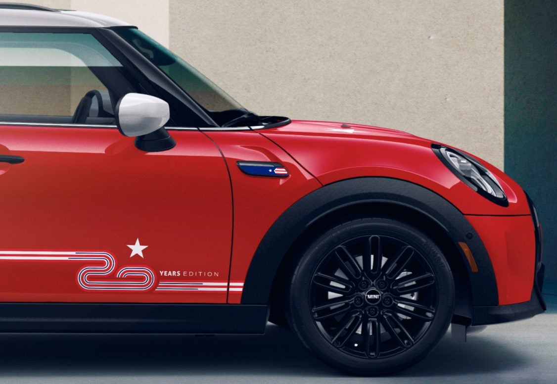 Vista lateral de un MINI 20 Years Edition Hardtop 4 Door Cooper S Chili Red aparcado sobre una superficie gris con un edificio beige al fondo. | MINI of Sterling en Sterling VA