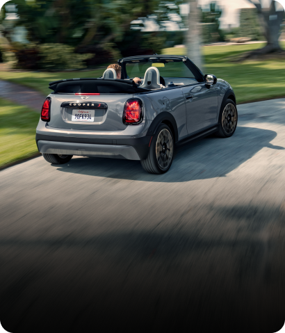 Vista trasera de un MINI Cooper S Descapotable en color Icy Sunshine Blue girando en una curva de la carretera con un edificio azul difuminado al fondo.