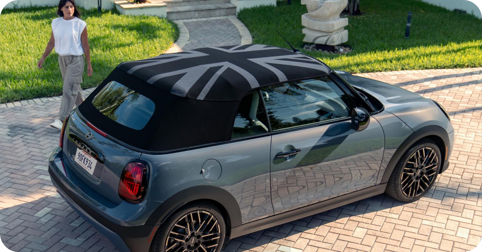 Mujer de pie detrás de la vista lateral de MINI Cooper Convertible gris en una calle suburbana.