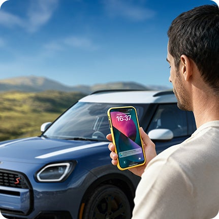 Una persona de pie junto a un MINI Countryman S ALL4, con un smartphone en la mano y mirando la pantalla. 