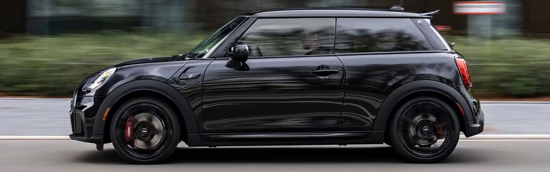 Vista lateral de un vehículo MINI John Cooper Works 1to6 Edition circulando solo por una calle mirando hacia la izquierda, con arbustos al fondo a lo largo y el entorno del vehículo difuminado.