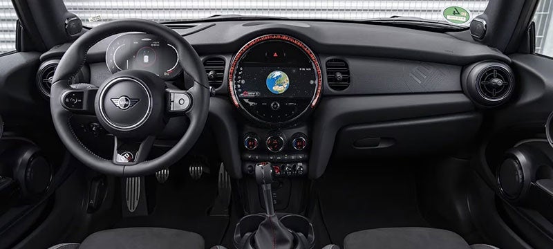 Primer plano de la fila delantera de un vehículo MINI John Cooper Works 1to6 Edition desde la perspectiva de la fila trasera, con un muro metálico y una valla metálica visibles a través del parabrisas en el fondo.