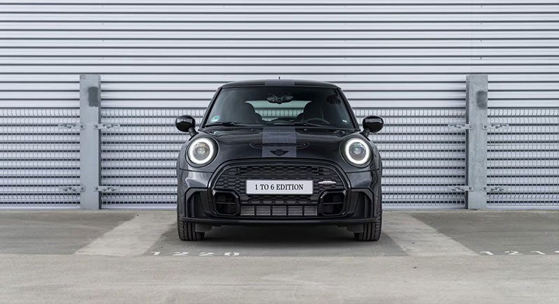 Vista frontal de un MINI John Cooper Works 1to6 Edition en color de carrocería Midnight Black II, aparcado en una superficie de acera con un muro metálico y una valla metálica detrás.
