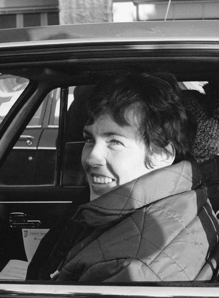 Pat Moss sentado en el asiento del conductor de un coche, en una foto en blanco y negro, sonriendo y mirando por la ventanilla.